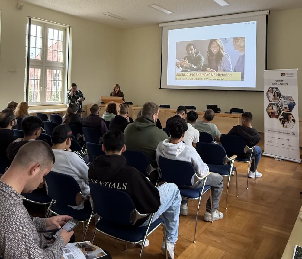 Teilnehmende sitzen im Vortragssaal vor einer Leinwand mit der Präsentation 'Ausbildungskampagne Migration – Sachsen-Anhalt will DICH!'