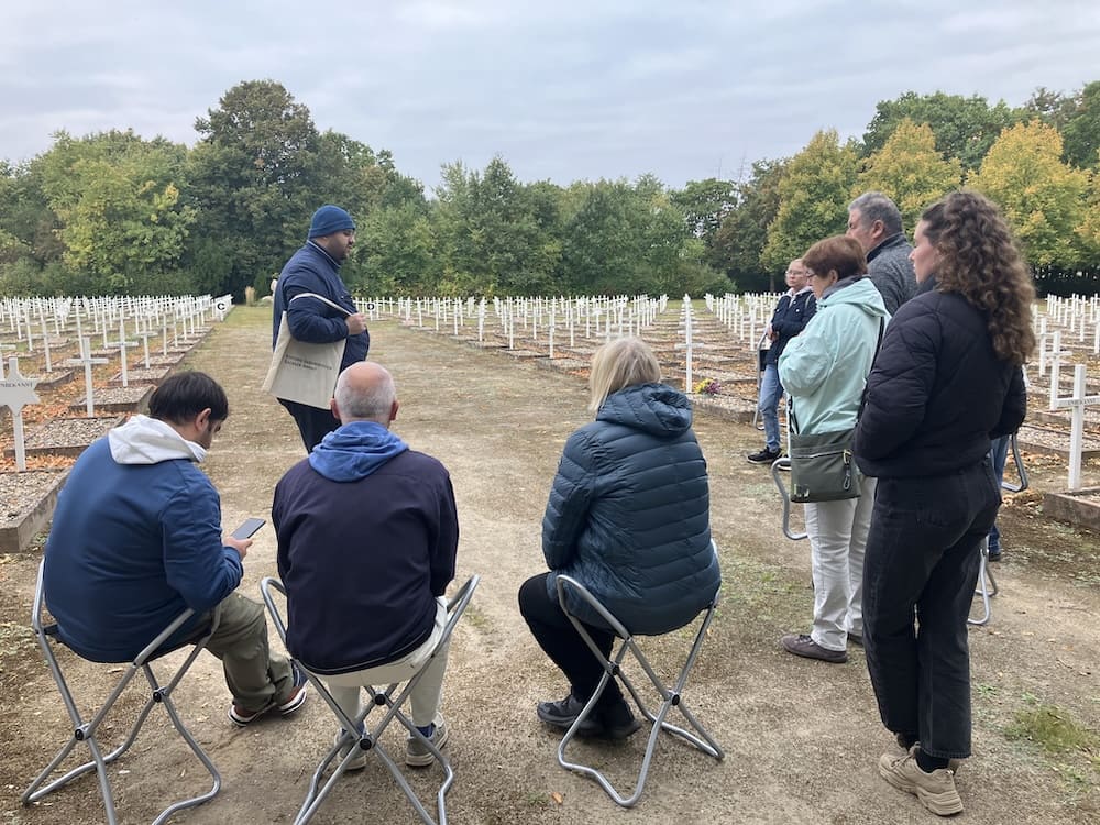 nem Guide zu, der zwischen Reihen weißer Kreuze auf einem Friedhof erklärt.