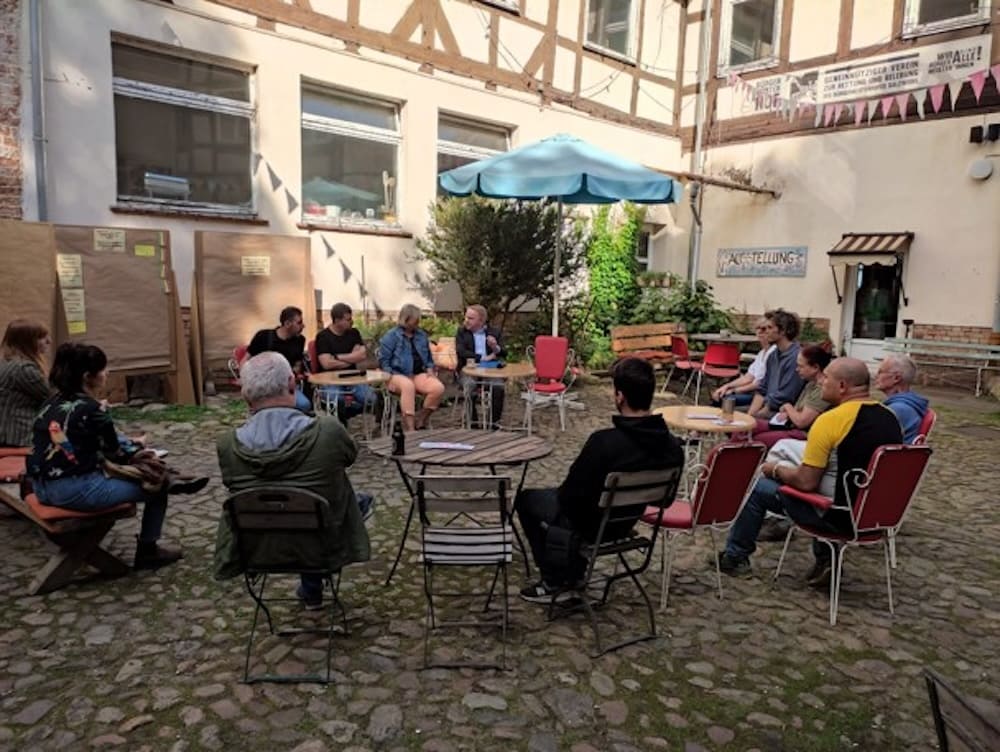 eilnehmende sitzen im lockeren Gesprächskreis in einem sonnigen Innenhof mit Sonnenschirm.